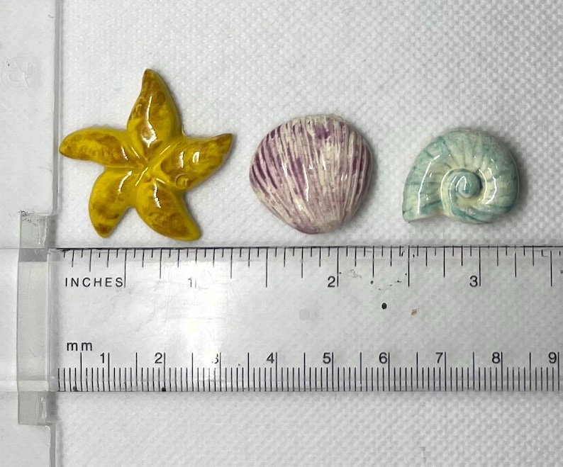 Puede incluir: Tres adornos de cer&aacute;mica con tem&aacute;tica marina: una estrella de mar amarilla, una concha p&uacute;rpura y blanca y una concha azul y blanca. Los adornos miden aproximadamente 2,5 cm.