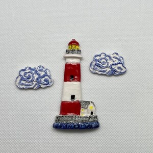 May include: A ceramic lighthouse figurine with a red and white striped tower, a yellow light, and a small white house at the base. The lighthouse is surrounded by two blue and white clouds.