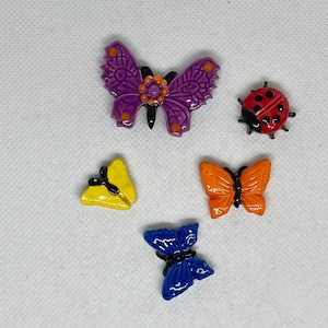 May include: A collection of colorful insect-themed decorative pins. The pins include a purple butterfly, a red ladybug, a yellow butterfly, an orange butterfly, and a blue butterfly. Each pin has a detailed design and is set against a white background.