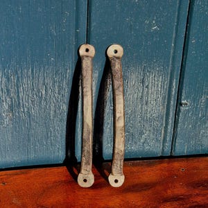 May include: A pair of aged metal door pulls. Each pull is rectangular with rounded ends and a hole for fixing. The metal has a worn, grey appearance, indicating age. The background is a blue wooden surface. The pulls are likely for a cabinet or door.