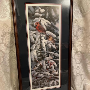May include: Framed print of a winter scene with cardinals and chickadees perched on a snow-covered pine tree. The birds are red, gray, and white, and the background is a soft blue.