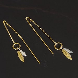 Puede incluir: Un par de pendientes de oro con una cadena delicada y un pequeño encanto circular con dos acentos de plumas. Una pluma es dorada y la otra es plateada.