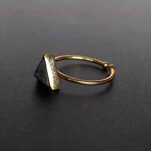 May include: A gold ring featuring a triangular, dark grey stone. The stone is set in a gold bezel. The ring band is thin and adjustable. The ring is set against a dark background.