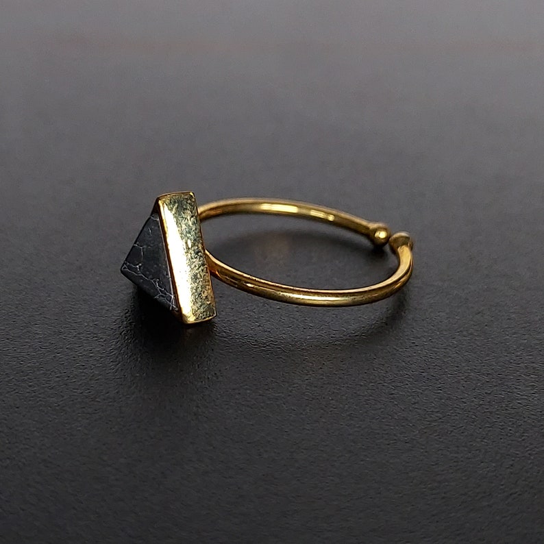 May include: A gold ring featuring a triangular, dark grey stone. The stone is set in a gold bezel. The ring band is thin and adjustable. The ring is set against a dark background.