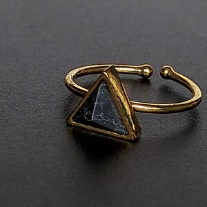 May include: A gold-coloured adjustable ring with a triangular, dark grey stone with white veining. The ring's band is open, with small ball-shaped details at the ends. The ring is set against a dark background.