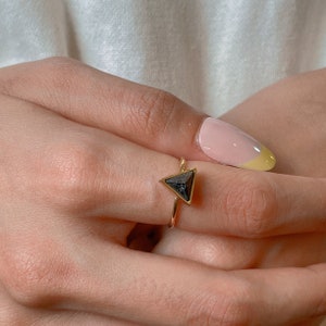 May include: A gold ring with a triangular, dark grey gemstone. The ring has a slender band and a minimalist design. The gemstone is set in a gold bezel, creating a contrast. The ring is a piece of jewellery.
