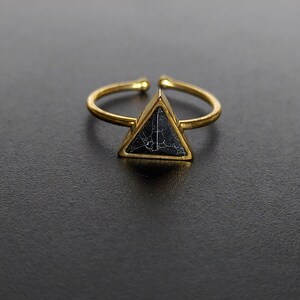 May include: A gold-tone adjustable ring with a triangular black stone, veined with white. The open band allows for size adjustment. The ring is set against a dark background, highlighting the jewellery.
