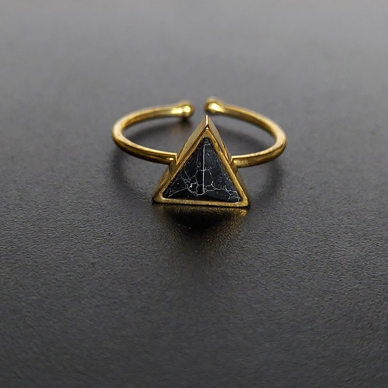 May include: A gold-tone adjustable ring with a triangular black stone, veined with white. The open band allows for size adjustment. The ring is set against a dark background, highlighting the jewellery.