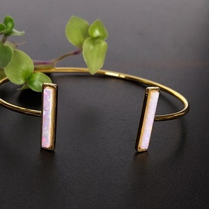 May include: A gold bangle bracelet with two rectangular opal stones. The stones are a pale pink color.