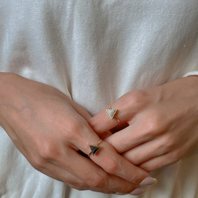 May include: Two gold rings, each with a triangular gemstone. One ring has a light grey stone, the other a dark grey stone. The rings are displayed against a soft white backdrop, highlighting the jewellery's design and colour.