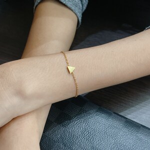 Può includere: Un delicato braccialetto color oro con un piccolo ciondolo a forma di triangolo. Il braccialetto ha un design a catena semplice e il triangolo è il punto focale. Il gioiello è indossato su un polso.