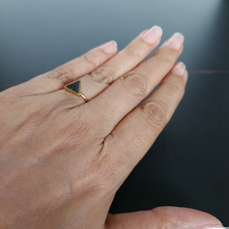 May include: A gold ring with a dark grey triangular stone. The ring has a thin band and a triangular stone at the top. The ring is displayed on a person's hand, set against a dark background.