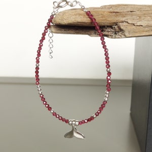 Op de afbeelding: Een rode edelsteen kralen armband met een zilveren walvisstaart bedel. De armband is gemaakt met kleine, gefacetteerde rode edelstenen en zilveren kralen.