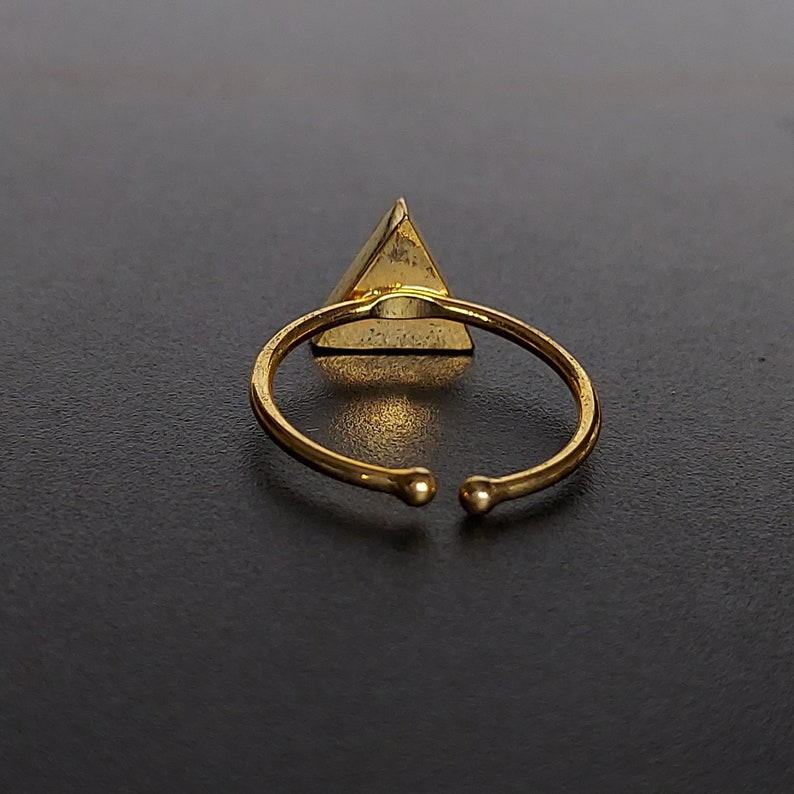 May include: Gold-coloured adjustable ring with a pyramid-shaped charm. The ring's band is open-ended with small ball accents. The pyramid charm has a textured surface and is centred on the ring.