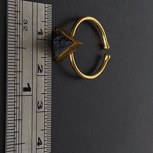 May include: A gold-coloured adjustable ring featuring a triangular, dark blue stone. The ring's band is open, allowing for size adjustment. The stone has a marbled appearance. The ring is set against a dark background.