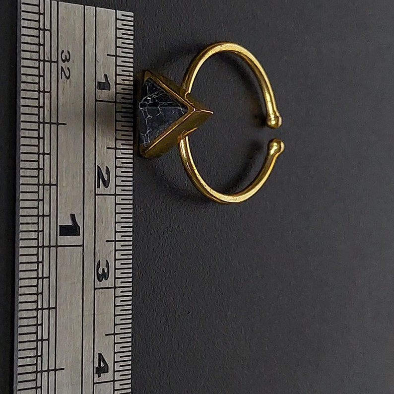 May include: A gold-coloured adjustable ring featuring a triangular, dark blue stone. The ring's band is open, allowing for size adjustment. The stone has a marbled appearance. The ring is set against a dark background.