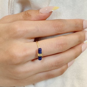 May include: A gold ring with a dark blue rectangular stone. The ring is on a person's finger.