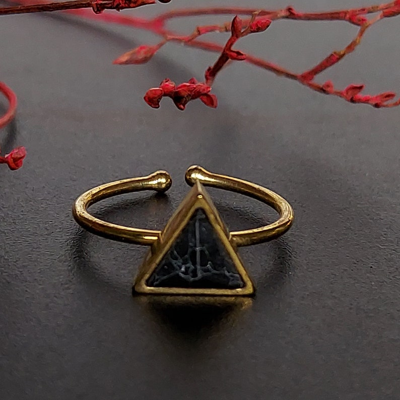 May include: An adjustable ring in gold colour, featuring a triangular, dark blue stone with a textured pattern. The ring's band is open at the top for size adjustment. The ring is set against a dark background with red floral accents.
