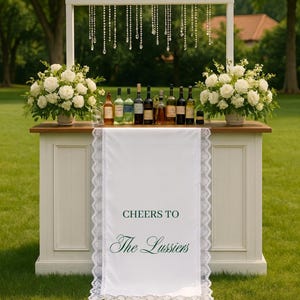 Pode incluir: Um bar branco com uma bancada de madeira, decorado com rosas brancas e garrafas de vinho. Uma faixa branca com o texto "Cheers to The Lussiers" está pendurada na frente. Fios de cristal pendem de uma moldura branca acima do bar.