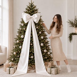 Puede incluir: Un gran lazo de cinta de satén color blanco roto con dos largas colas que caen por la parte delantera de un árbol de Navidad verde decorado con luces blancas cálidas y adornos redondos, con regalos envueltos en la base.