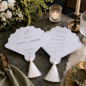 May include: Two white shell-shaped wedding programmes with scalloped edges and white tassels. The left programme features the text 'Welcome to the wedding of Oliva & Jeremy' and the date '09.18.2027'. The right programme displays a wedding schedule.