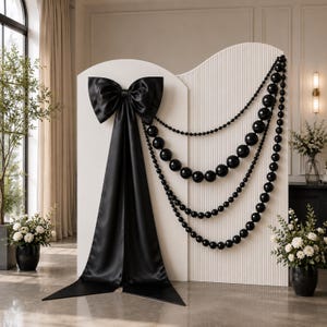 Peut inclure: Un paravent blanc arqué et cannelé, orné d'un grand nœud en satin noir et de plusieurs rangées de perles noires brillantes de tailles variées. Le paravent présente un bord supérieur incurvé et une texture nervurée verticale sur toute la surface.