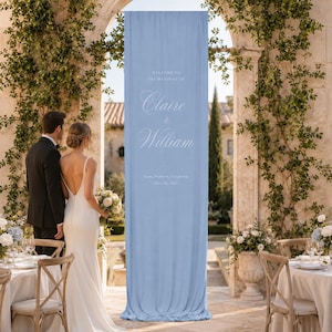 May include: A tall, light blue fabric wedding welcome sign featuring white text that reads: Welcome to the wedding of Claire & William, Santa Barbara, California, June 14, 2024. The banner hangs vertically within a stone archway.