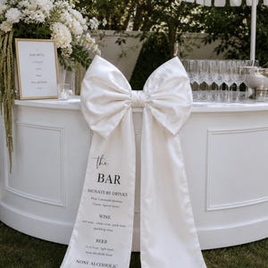 May include: A white satin fabric sign featuring a large decorative bow at the top. The text reads: the BAR, SIGNATURE DRINKS, Pink Lemonade Mimosa, Classic Aperol Spritz, WINE, red wine, white wine, pink wine, sparkling wine, BEER, dark beer, light beer, none alcoholic beer, NONE ALCOHOLIC, soft drinks, orange juice, sparkling water.