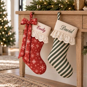 Könnte beinhalten: Zwei Weihnachtsstrümpfe hängen an einem Holztisch. Der linke Strumpf ist rot mit weißen Schneeflocken und einer großen Schleife, personalisiert mit dem Namen Emily. Der rechte Strumpf hat grüne und weiße Streifen und ist mit dem Namen David versehen.