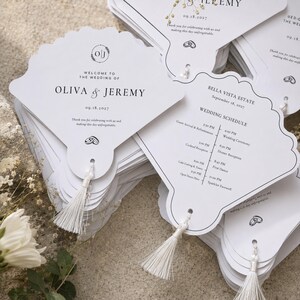 May include: A stack of white, fan-shaped wedding programmes with scalloped edges and white tassels. The front features the text 'Welcome to the wedding of Oliva & Jeremy 09.18.2027'. The back displays a wedding schedule for Bella Vista Estate.