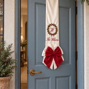 May include: A long, cream-coloured fabric door banner hanging vertically. It features an embroidered green holly wreath with a red bow, a red letter W in the centre, and the red embroidered text The Wilsons. A large, deep red fabric bow is at the base.