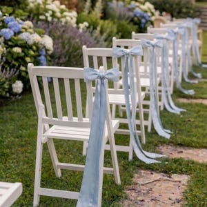 Op de afbeelding: Een rij witte houten klapstoelen, elk versierd met lichtblauwe satijnen strikken en linten. De stoelen staan op een grasveld, met bloemen op de achtergrond, wat duidt op een buitenevenement.