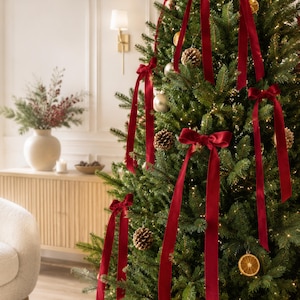 May include: A lush green Christmas tree decorated with long, deep red velvet ribbons tied into bows. The tree features natural pinecones, dried orange slices, and metallic silver ornaments nestled among the branches, illuminated by warm white string lights.