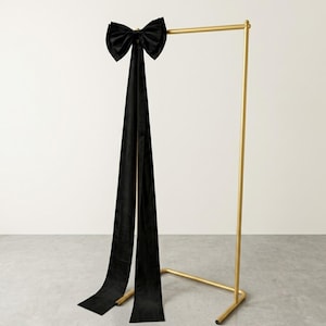 May include: A black velvet bow with long, flowing tails is displayed on a gold metal clothing rack. The bow is tied at the top of the rack, and the tails hang down to the floor. The rack has a simple, minimalist design.