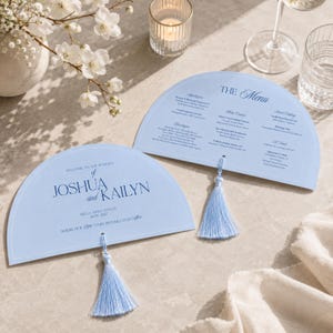 May include: Two semi-circular light blue paper cards featuring dark blue text. One card displays the names Joshua and Kailyn with wedding details, while the other lists a menu. Each card includes a matching light blue tassel attached at the base.