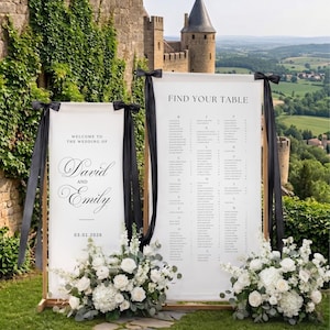 May include: Two white wedding signs with black text and black ribbon accents. One sign reads "Welcome to the Wedding of David and Emily" and the date. The other sign says "Find Your Table" with guest names. White floral arrangements are at the base.