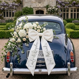 Peut inclure: Une voiture vintage bleu marine décorée pour un mariage. Un grand nœud blanc est attaché à la voiture, avec les mots "Emily & Hayden" et "Just Married" écrits sur les rubans. Des fleurs blanches et de la verdure ornent la voiture.