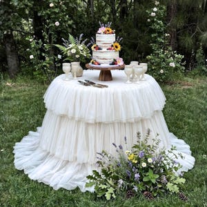 May include: A three-tiered white cake adorned with sunflowers, berries, and flowers on a wooden stand. The cake is on a round table draped with white, ruffled fabric. The table is set outdoors with glasses and cutlery.