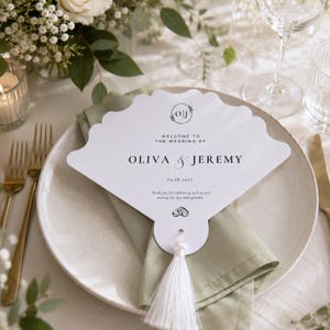 May include: A white, scalloped, fan-shaped wedding programme card featuring the text "Welcome to the wedding of Oliva & Jeremy," the date "09.18.2027," a monogram "O|J," and a small illustration of two wedding rings, finished with a white tassel.