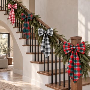 May include: Four decorative fabric bows attached to a green pine garland draped along a wooden staircase railing. The bows feature distinct patterns including red and white gingham, dark green plaid, black and white buffalo check, and red and green tartan.