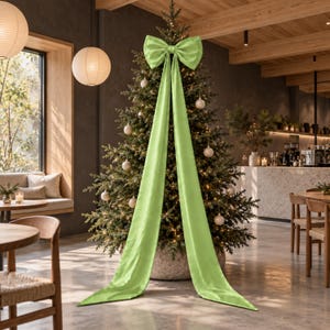 Peut inclure: Un grand nœud en tissu vert citron vif avec deux longs rubans tombants drapés sur le devant d'un sapin de Noël décoré. L'arbre est orné de petites boules de Noël de couleur neutre et de guirlandes lumineuses blanc chaud.