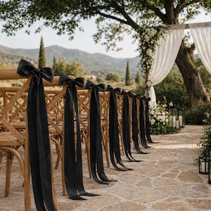 Puede incluir: Sillas de madera con lazos de satén negro y largas cintas dispuestas en filas para un evento al aire libre. Las sillas miran hacia un arco cubierto con tela blanca y arreglos florales. El escenario es un camino de piedra con un telón de fondo montañoso.