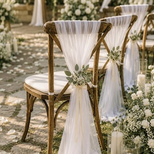 Op de afbeelding: Houten stoelen met witte kussens en transparante stof over de ruggen gedrapeerd. Elke stoel is versierd met een klein bloemstuk en lint. De setting suggereert een buitenevenement of bruiloft.