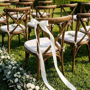 Peut inclure: Rangée de chaises en bois avec coussins crème sur une pelouse. Des rubans blancs sont attachés aux chaises, et un arrangement floral de roses blanches et de verdure est au premier plan. Les chaises ont un dossier en forme de X.