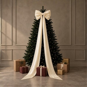 Puede incluir: Un gran lazo de tela color crema con dos largas cintas fluidas que caen por la parte delantera de un árbol de Navidad verde oscuro. El árbol está iluminado con pequeñas luces y rodeado de varias cajas de regalo en tonos neutros.