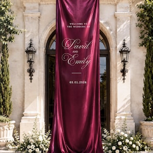 Op de afbeelding: Een lange, verticale banner van diepbordeauxrode satijnstof met witte bedrukte tekst: WELCOME TO THE WEDDING OF David AND Emily 03.01.2026. De banner hangt recht naar beneden en vormt onderaan een lichte plooi op de grond.