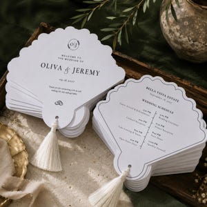 May include: Two stacks of white, shell-shaped wedding program fans with scalloped edges. One stack displays the names Oliva & Jeremy with the date 09.18.2027, while the other shows a wedding schedule. Each fan features a cream-colored tassel.