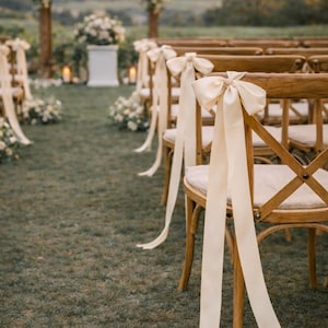 Puede incluir: Escena de ceremonia de boda al aire libre con sillas de madera adornadas con lazos y cintas color crema. Las sillas están dispuestas en un césped, que conduce a un arco floral y un altar.