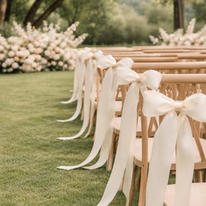 Puede incluir: Sillas de madera con lazos y cintas color crema alineadas en un pasillo de césped. Las sillas están dispuestas en filas, con los lazos atados a los respaldos. Flores blancas y árboles en el fondo crean un ambiente sereno.