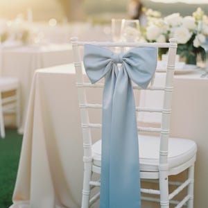 Puede incluir: Una silla de madera blanca con un lazo de satén azul claro atado a la parte posterior. Las largas colas del lazo caen por la silla. La silla está preparada para un evento al aire libre, con una mesa y flores al fondo.